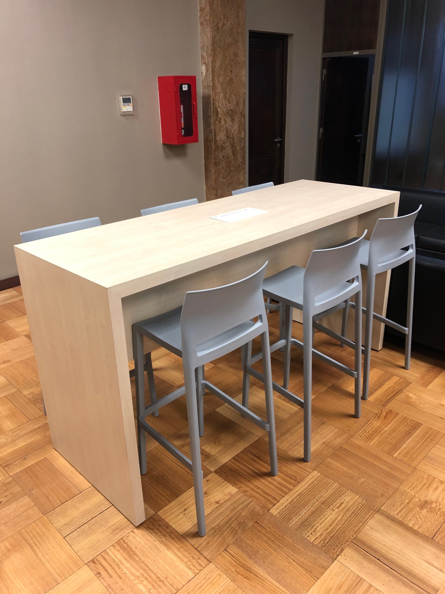 Mesa alta de madera clara con taburetes grises en un espacio compartido
